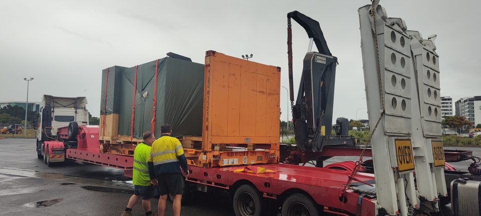 Two workers inspect oversized cargo on a flat rack container, prepared for delivery using a dual-truck setup in Brisbane.